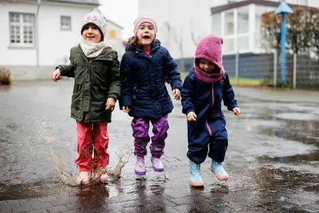 Ev. Kindertagesstätte "Kinder Garten Eden" Gemünden Drei Kinder in bunter Regenkleidung und Gummistiefeln spielen im Regen - treten in Pfützen. Dahinter sind Gebäude der KiTa zu sehen.